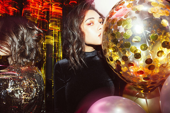 Woman celebrating with fancy glasses at a party, glamorous setting with colorful balloons and reflections.