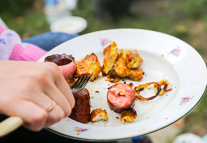 A person's hand holding a fork near a plate of leftovers, including grilled sausage and vegetables, at an outdoor gathering.
