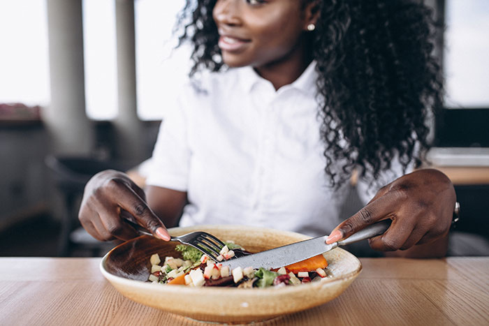 Woman in a white shirt eating a salad with a knife and fork at a wooden table, focused on her meal. Woman in a white shirt eating a salad with a knife and fork at a wooden table, focused on her meal.