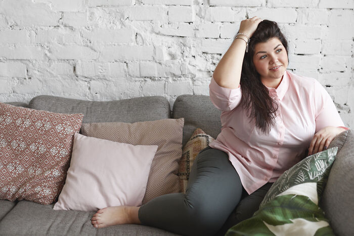 Woman in a pink shirt sitting on a gray sofa, surrounded by cushions, touching her hair.