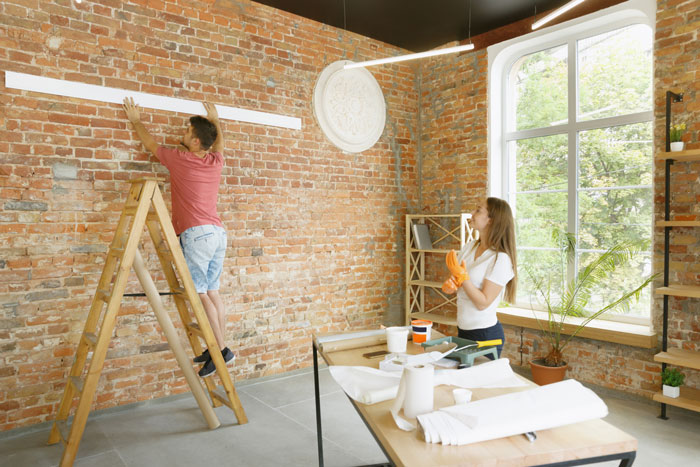 A couple involved in property remodel, with the man on a ladder in a brick-walled room.