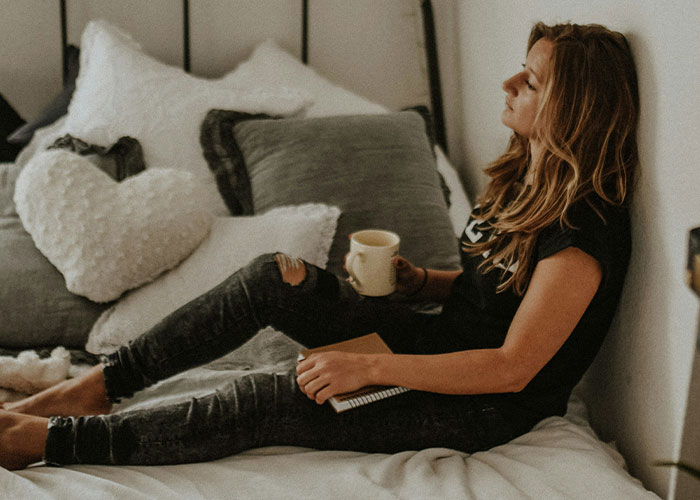 Woman sitting on bed with a mug, deep in thought, symbolizing emotional distance caused by an insidious work wife.