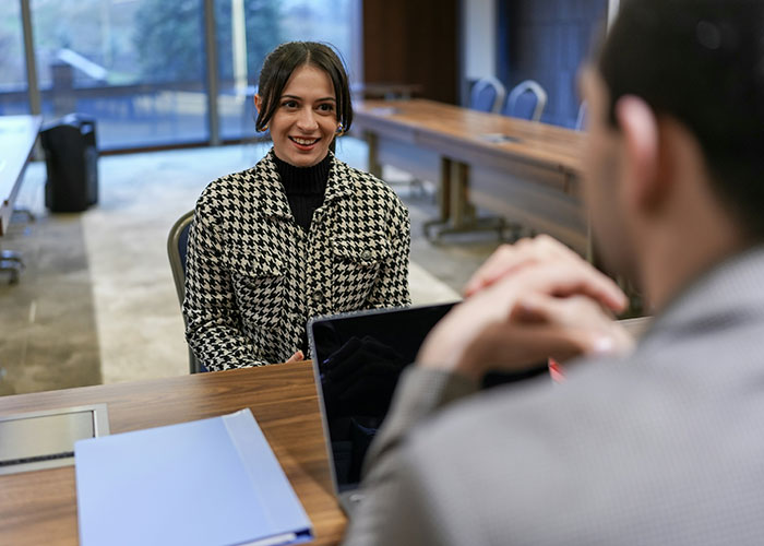 A woman in an interview, wearing a houndstooth coat, smiling across a desk with a folder, symbolizing a workplace moment.