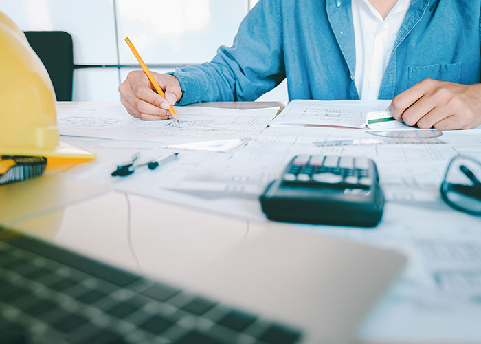 Person working on blueprints, blue shirt, toxic workplace concept, hard hat and calculator on desk.