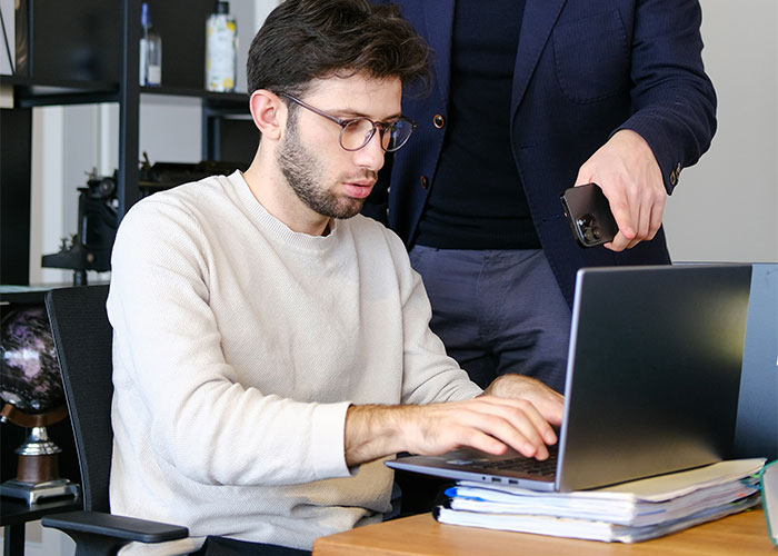 Man in an office working on a laptop with another person pointing at the screen, illustrating a toxic workplace scenario.