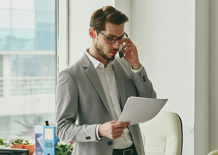 Man in an office, wearing a gray suit and glasses, reviews documents by a window, illustrating workplace dynamics.