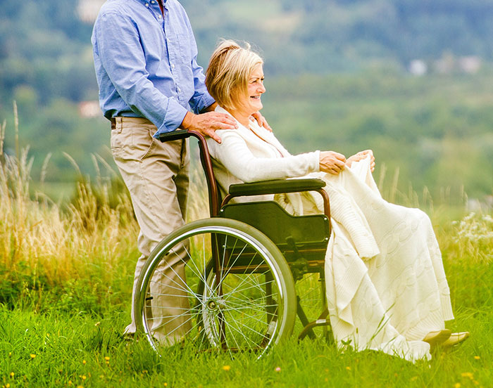 A person stands behind a woman in a wheelchair, both smiling, in a scenic outdoor setting.