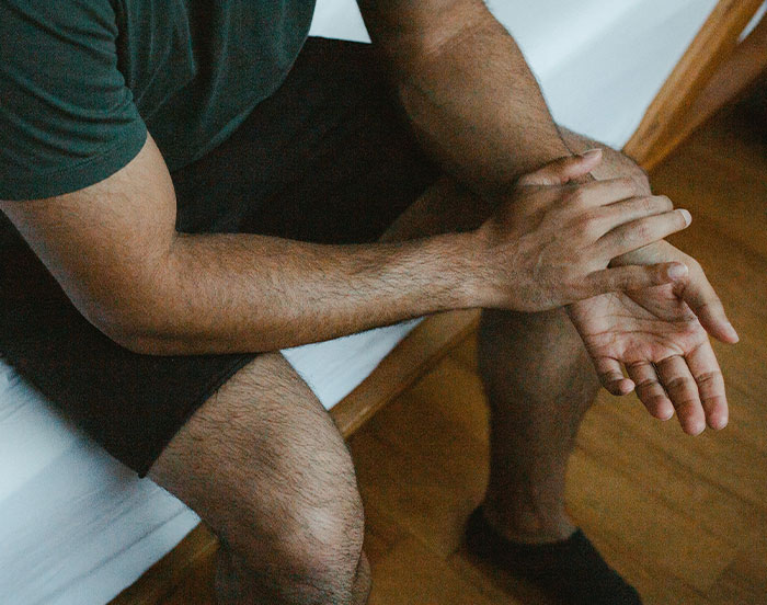 Anxious person sitting on a bed, hands clasped, wearing a green shirt and black shorts, possibly seeking attention.