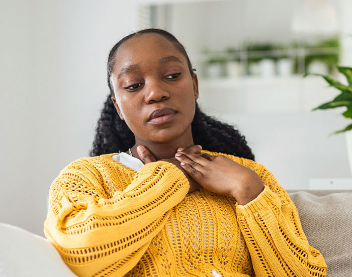 Person in a yellow sweater, sitting with a thoughtful expression, hands near neck, indoors.