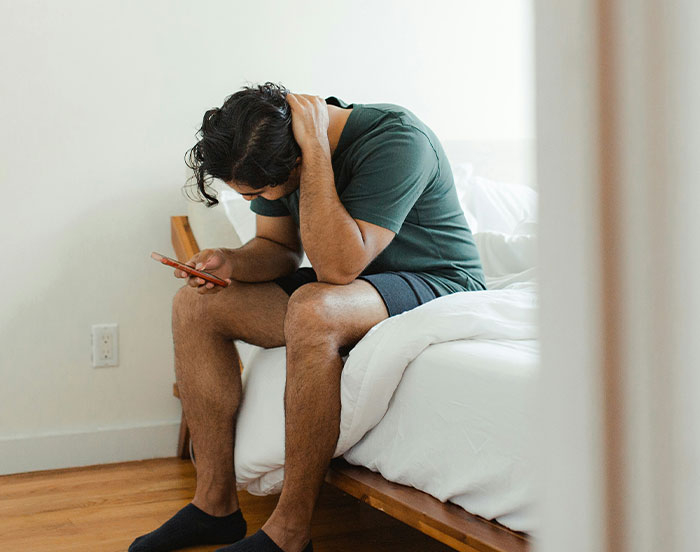 "Man in green shirt sitting on bed, looking at phone, appearing thoughtful."