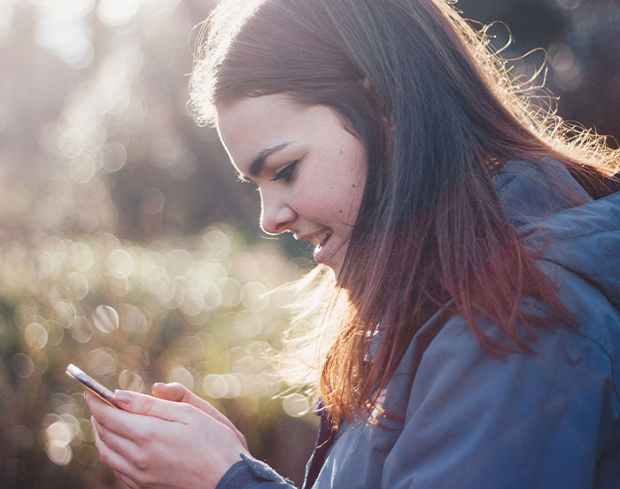 Young woman in a park, wearing a blue jacket, looking at her phone, smiling in natural light.