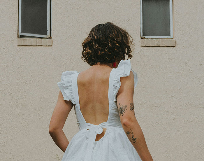 Person in a white dress stands against a wall, displaying tattoos and curly hair, seeking attention.