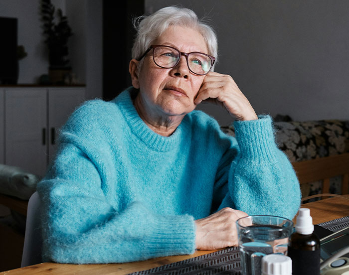 Elderly person in a blue sweater, sitting at a table with a thoughtful expression, seeking attention.