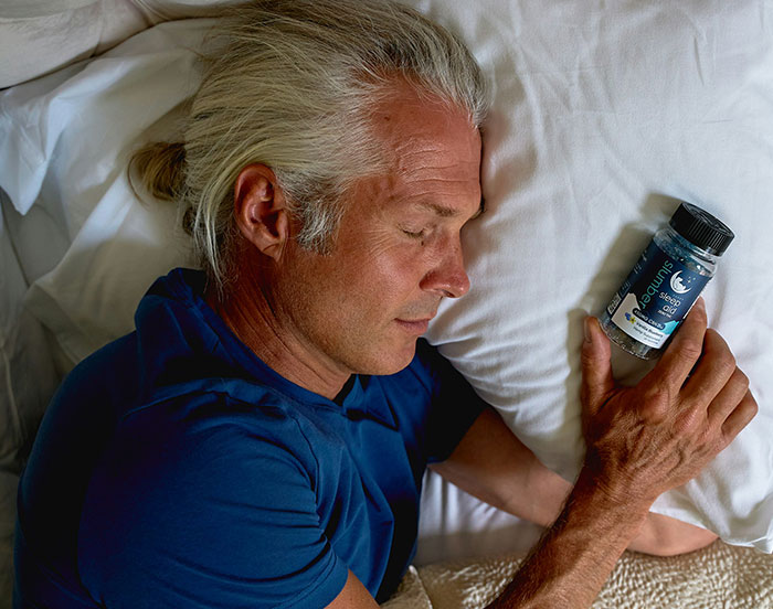 Man in blue shirt holding a supplement bottle, resting on a pillow, showcasing unhinged attention-seeking behavior.