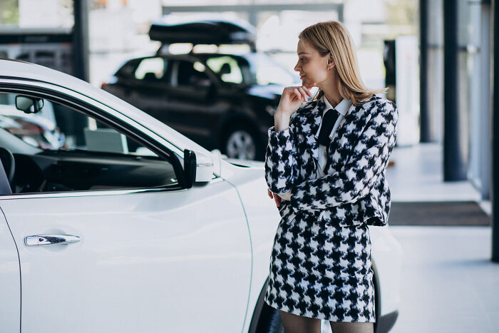 Woman examining a white car in a showroom, wearing a houndstooth suit, highlighting common offensive remarks.