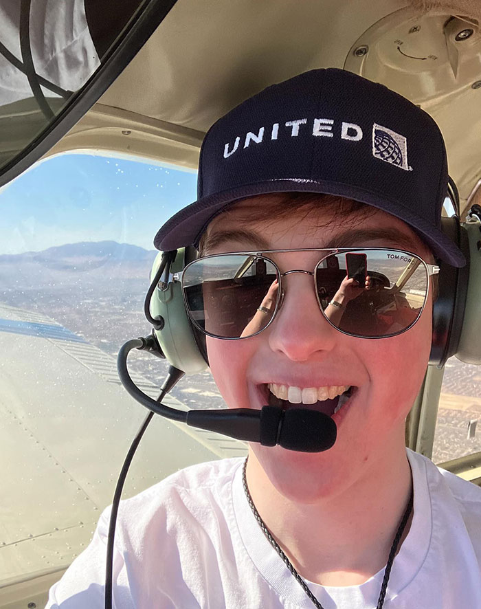 Young Sheldon cast member Iain Armitage flying a plane, wearing a headset and sunglasses, with a view of the landscape through the window.