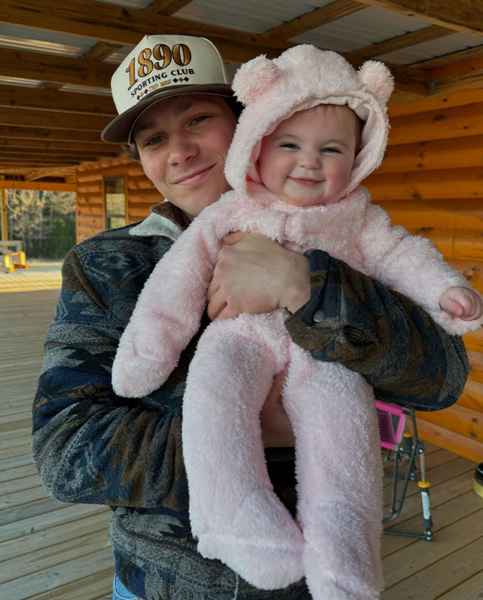 Montana Jordan holding a smiling baby in a pink fuzzy suit outside a log cabin, linked to Young Sheldon cast post-show activities.