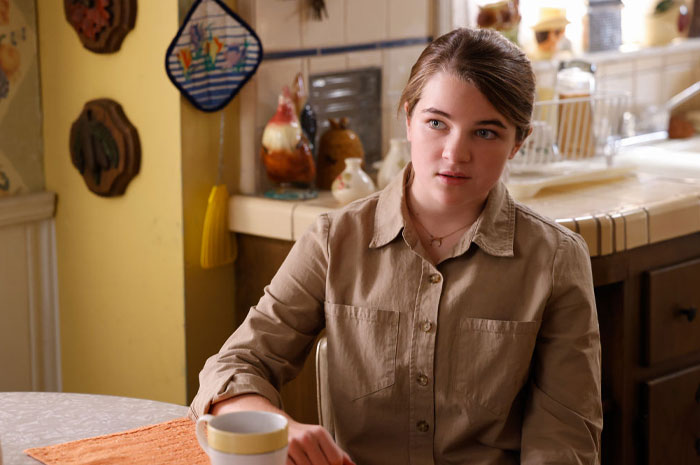 Young Sheldon cast member Raegan Revord sitting at a kitchen table, wearing a beige shirt, with a mug in front of her.