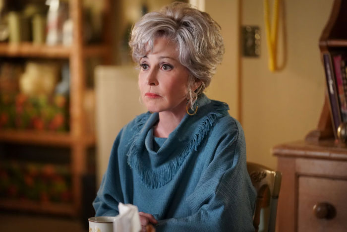 Actress Annie Potts from Young Sheldon cast, sitting in a cozy room, wearing a blue sweater and earrings, holding a cup.