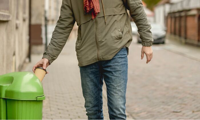 Person throwing a paper cup into a green trash bin on a street, representing everyday habits and cleanliness concerns.