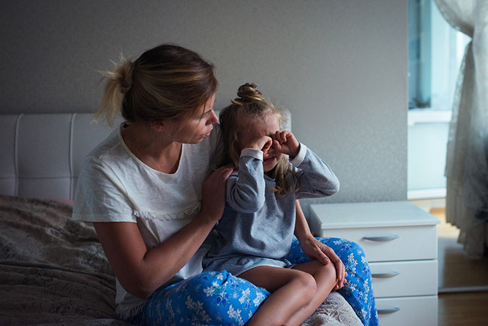 Woman comforting her 7-year-old child at home, excluded from spa day with friends.