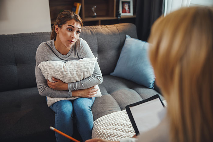 Woman on a couch, clutching a pillow, discussing adoption with another person in a home setting.