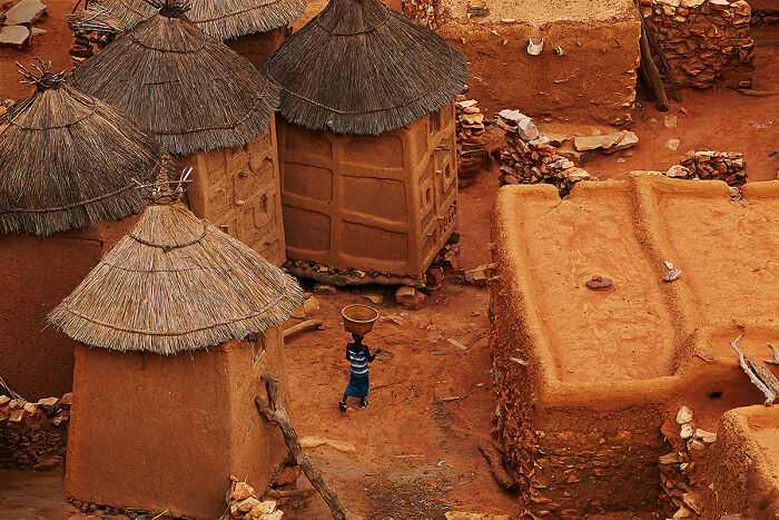 Aerial view of traditional huts and a person carrying a basket in a vibrant cultural village setting.