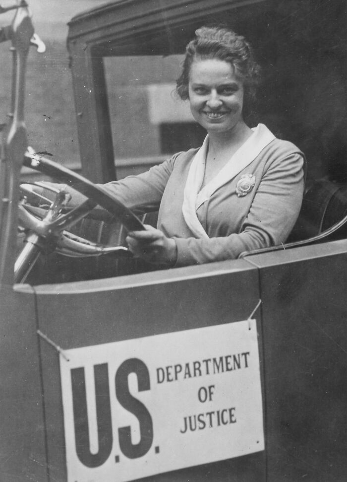 Smiling woman in vintage car with U.S. Department of Justice sign, showcasing rare historical moment.