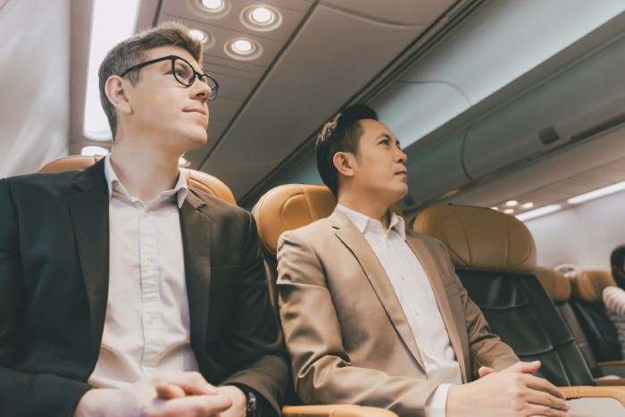 Two men in business attire sitting on a plane, discussing seat arrangements. Two men in business attire sitting on a plane, discussing seat arrangements.