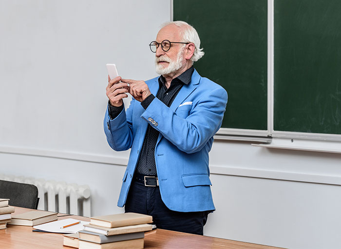 Professor in a blue blazer using a phone in a classroom, with books and a chalkboard in the background.