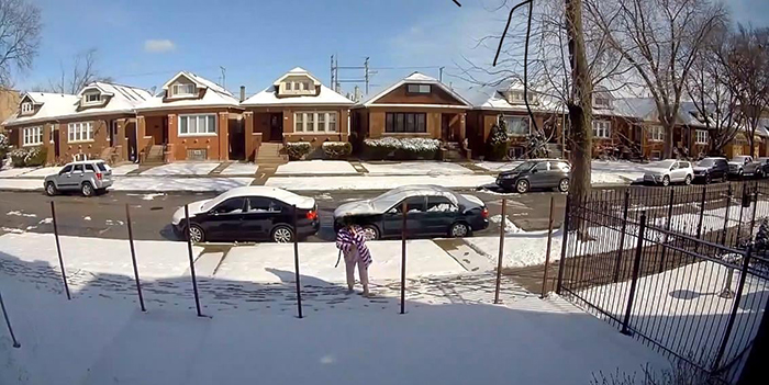 Woman on snowy street beside cars and houses. She appears to be examining or interacting with metal posts, possibly related to a fence.