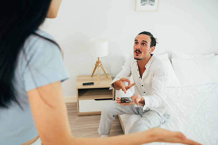 Man surprised about wife's secret monitoring, holding phone in bedroom discussion.