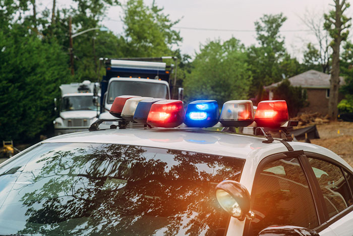 Police car with lights on in a suburban neighborhood, trees and houses visible in the background.