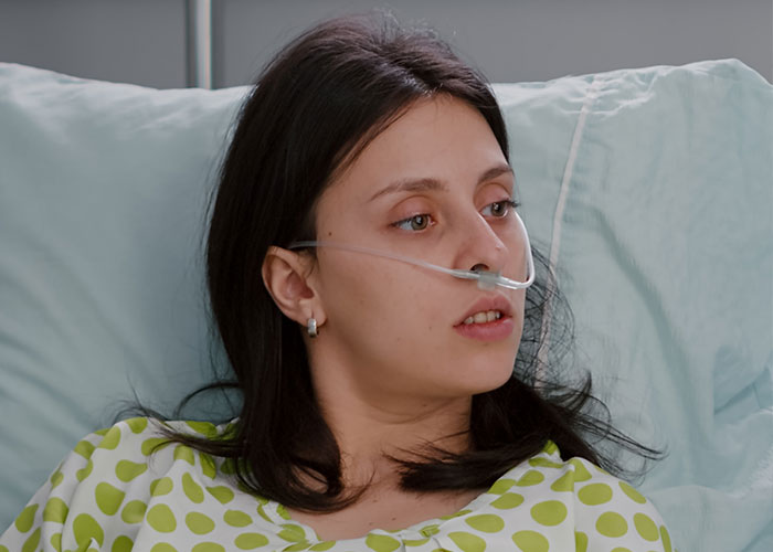 Woman in hospital room with nasal cannula, wearing a polka dot gown, lying in bed.