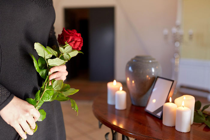 Person holding a rose beside a table with an urn and candles, symbolizing conflict over ashes with a hateful wife. Person holding a rose beside a table with an urn and candles, symbolizing conflict over ashes with a hateful wife.
