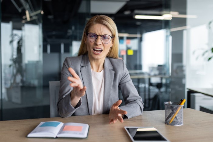 Woman in office gesturing emphatically, wearing a gray suit and glasses, representing an egoistic boss.