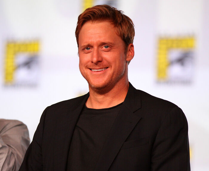 A smiling celebrity at a public event, wearing a black jacket, with a colorful background.