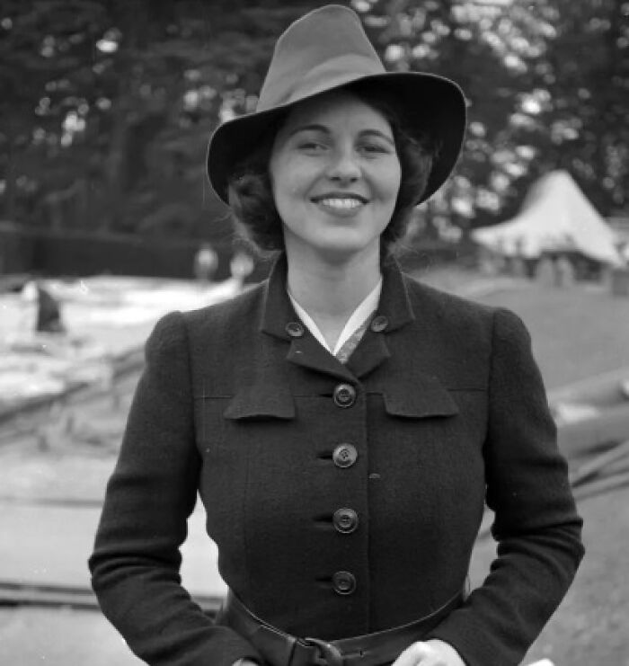 A woman in vintage attire and hat, smiling outdoors, representing historical fashion from the past.