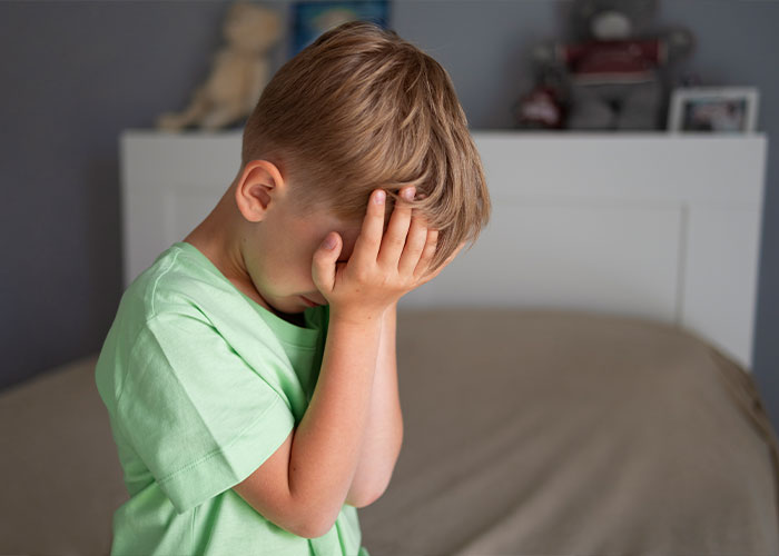 A young child in distress, covering their face in a bedroom, highlighting parenting challenges.