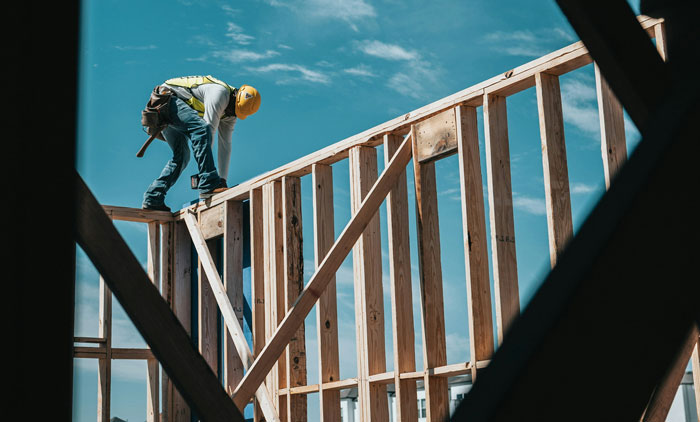 Construction worker in safety gear balancing on wooden framework under blue sky.