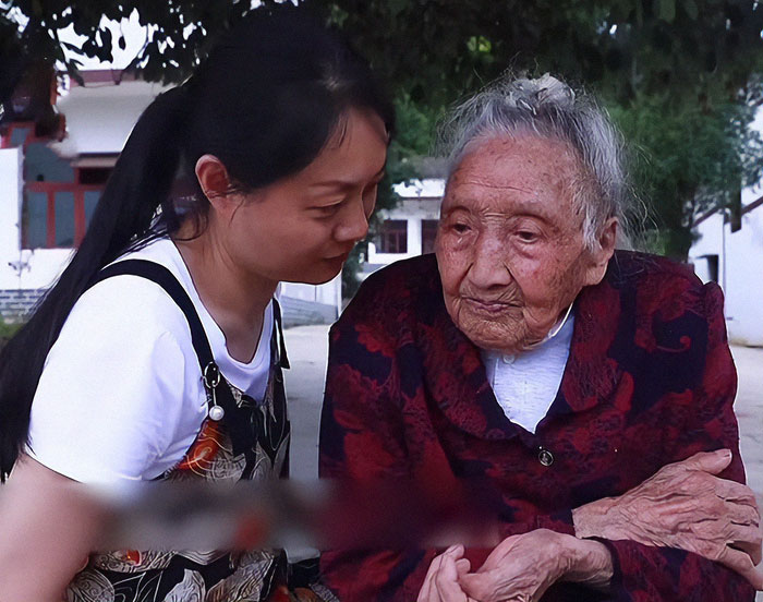 Elderly Chinese woman, 103, sitting outdoors with a young woman, wearing a red and black jacket, looking pensive.