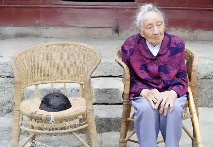 Elderly Chinese woman sitting outside on a wicker chair, wearing a purple jacket, with an empty chair beside her.