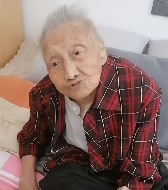 Elderly Chinese woman in a red plaid shirt, sitting upright in a chair, reflecting a lifetime of patience and endurance.