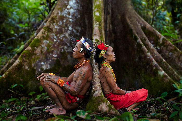 Two individuals in traditional attire sit back-to-back against a large tree, documenting culture and tradition.