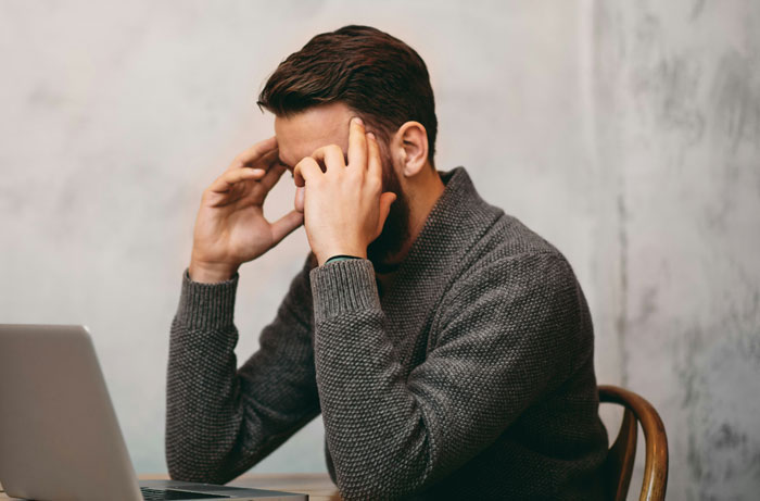 Man in a gray sweater sitting at a desk, stressed and holding his head, illustrating mental health issues.