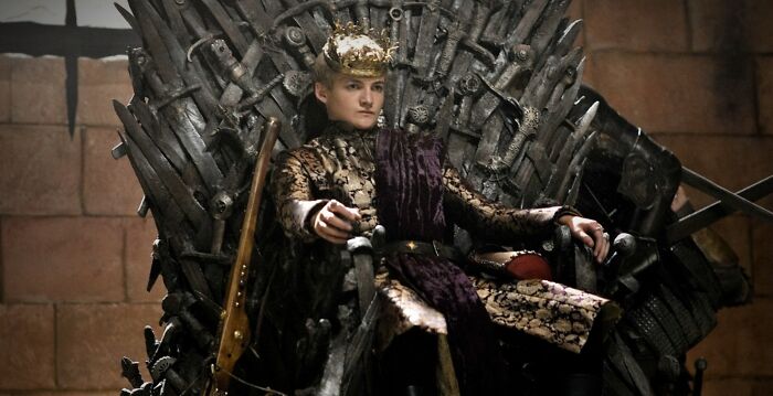 Actor sitting on an iron throne, wearing a crown and regal attire, embodying a role he was born to play.