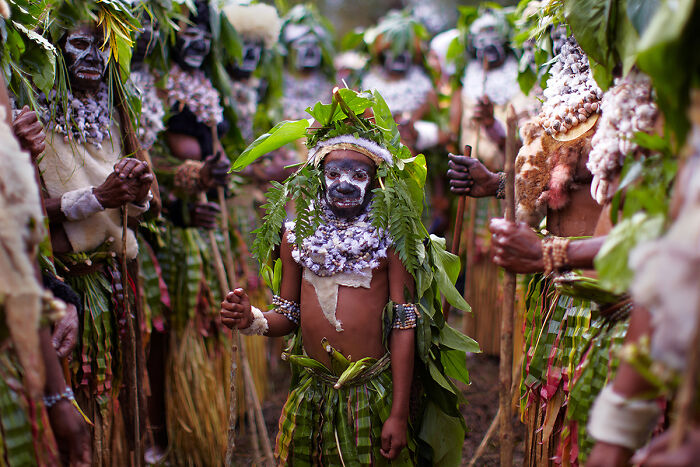 A child in traditional attire with painted face and leaves, surrounded by a group showcasing global culture and tradition.