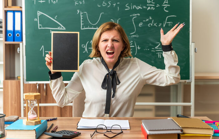 Teacher in a classroom holds a small chalkboard with an exasperated expression, illustrating work revenge.