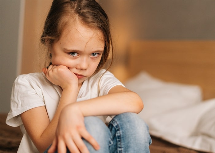 Sad child sitting on a bed, wearing casual clothes, reflecting parenting mistakes.