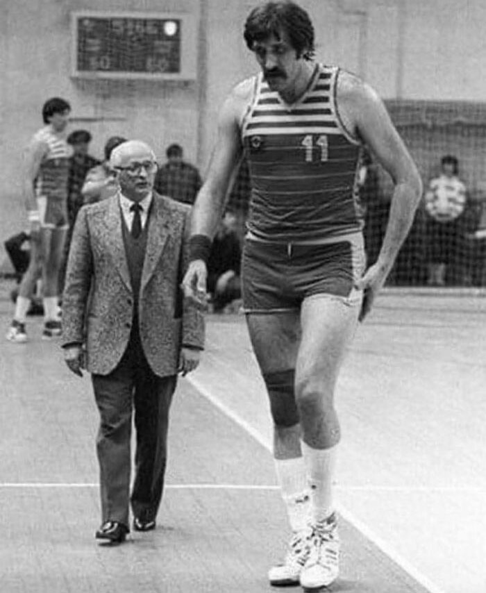 A very tall basketball player in uniform stands next to a shorter man in a suit, showcasing a striking height contrast.